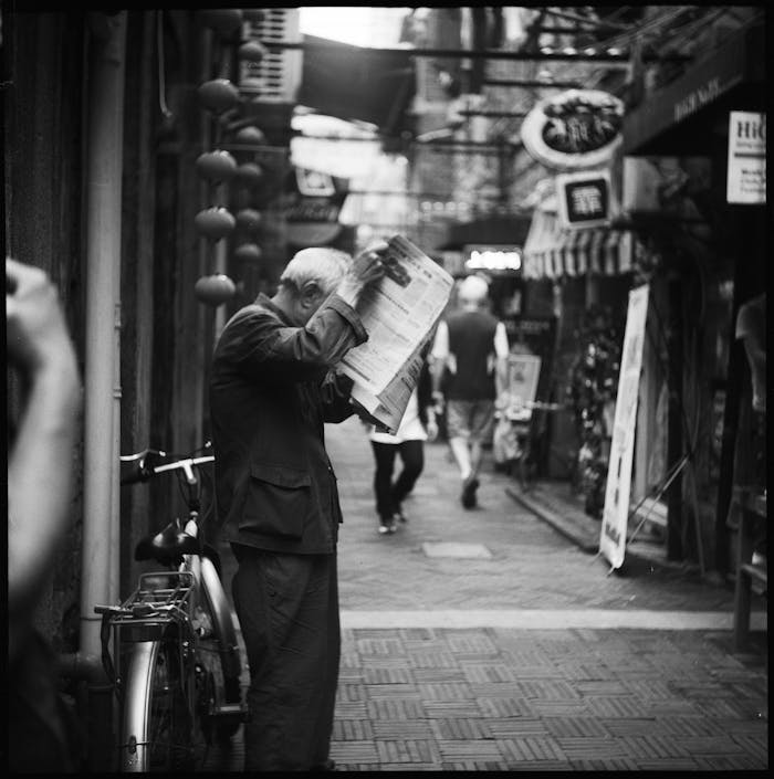 home-hero Elderly man reading a newspaper in a narrow city alleyway, a candid black and white street scene.